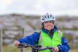 Smiling mature female cyclist with a blue jacket, helmet and anti-reflective vest next to a bicycle with a blurred background, relaxing day in Ireland