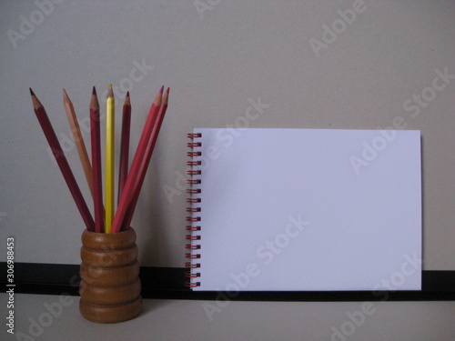 lápices de colores en gamas de rojos, rosas y amarillo en porta lápices marrón con cuaderno de anillas rojas con fondo de carpeta gris y negro. Lápices para dibujar y hacer bocetos. photo