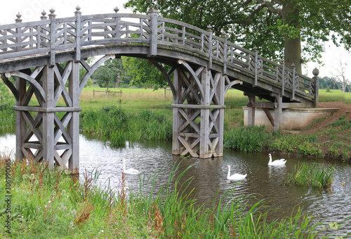 A wooden bridge in the countryside with three swans swimming underneath.