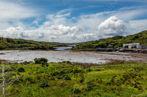 Clifden bay at low tide with boats anchored on the pier, surrounded by green vegetation on a sunny spring day with a blue sky and abundant white clouds in Clifden, Ireland