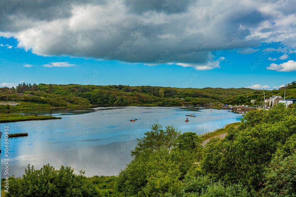 Clifden bay and its harbour at high tide with boats anchored in the water, seen from a hill, surrounded by trees and green vegetation, spring day with a blue sky and white clouds in Clifden, Ireland