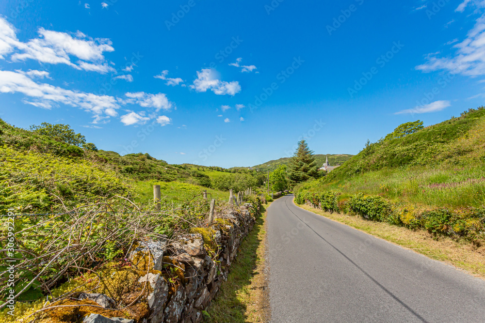 Rural road with limestone fences between hills with green vegetation, sunny spring day with a blue sky and white clouds in the province of Connacht, Ireland Rural road with limestone fences between hills with green vegetation, sunny spring day with a blue sky and white clouds in the province of Connacht, Ireland