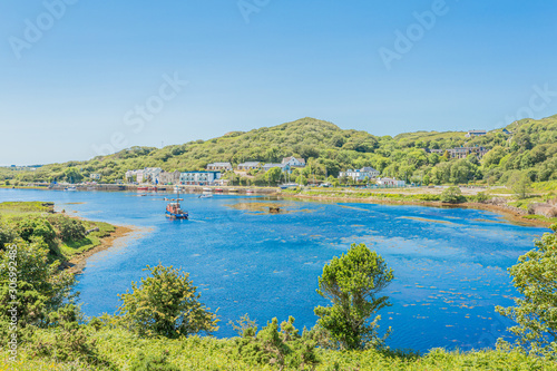 Hill with green vegetation, the port of Clifden at high tide and a boat in the background, surrounded by trees, sunny spring day with a blue sky in Clifden, Ireland