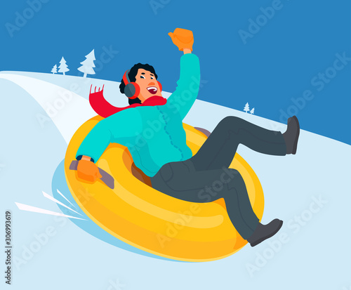 Smiling man in a down jacket, scarf, headphones going down a hill on an inflatable snow tube. Winter entertainment, sport, activity. 