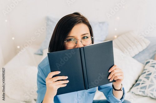 Young woman in glasses with black short hair covered her face with a book