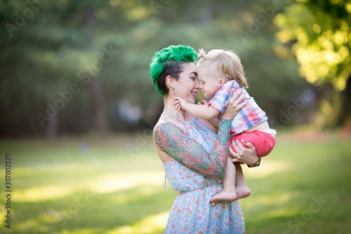 A portrait of Caucasian young woman with green hair and tattoo holding a blond toddler boy in her hands. Hugging, Mother's day concept