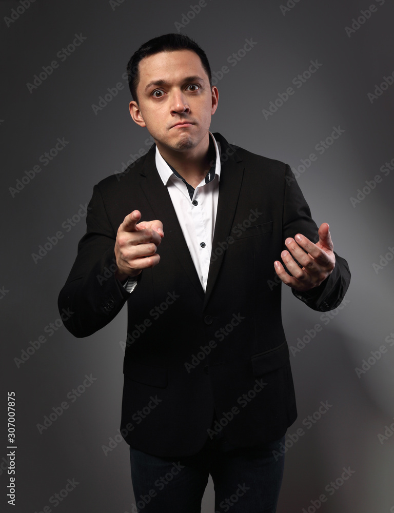 Surprising grimacing man looking in black suit with funny face showing the finger on grey studio background. Closeup Surprising grimacing man looking in black suit with funny face showing the finger on grey studio background. Closeup
