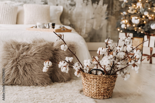 Dry Cotton Flowers in Wicked Basket, Woven Pillow on Carpet. Tray on Bed, Christmas Pine Tree Decorated Glowing Lamps, Toys and Presents and Grunge Wall on Blurred Background. Bedroom Cozy Interior