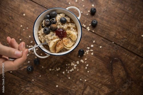 Vegetarian breakfast, woman's hand and porridge top view on wooden table with copyspace. Ingredients: oat flakes, banana, blueberries, chia, cinnamon, maple syrup and strawberry jam. Horizontal.