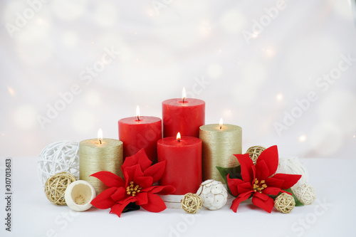 Red and gold Christmas candles surrounded by red poinsettia flowers, gold twig balls and natural potpourri elements on a white draped fabric background with white Christmas lights and bokeh effect.