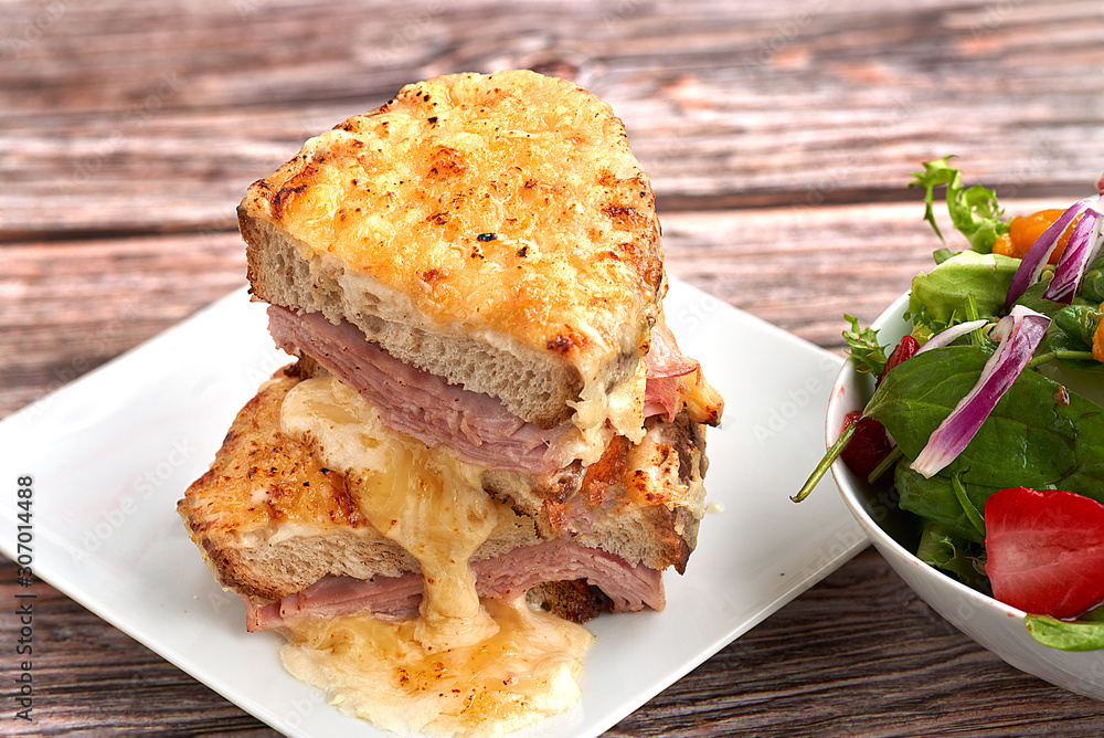 Sandwich with Parmesan and Gruyere cheese, ham, béchamel sauce and artisan bread accompanied by a salad on a wooden background.  The Croque Monsieur.