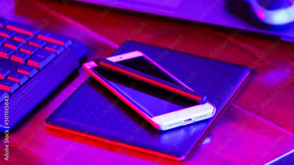 Freelancer workplace in neon light. Computer, graphics tablet and smartphone on the table. Place for creativity in blue pink light. Widescreen Freelancer workplace in neon light. Computer, graphics tablet and smartphone on the table. Place for creativity in blue pink light. Widescreen