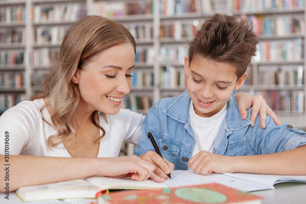 Close up of a cheerful beautiful wman helping her son with school assignment at the library Close up of a cheerful beautiful wman helping her son with school assignment at the library