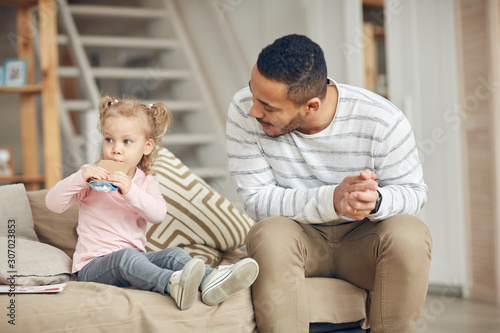 Portrait of contemporary dad looking at cute little daughter drinking juice while sitting on sofa at hone