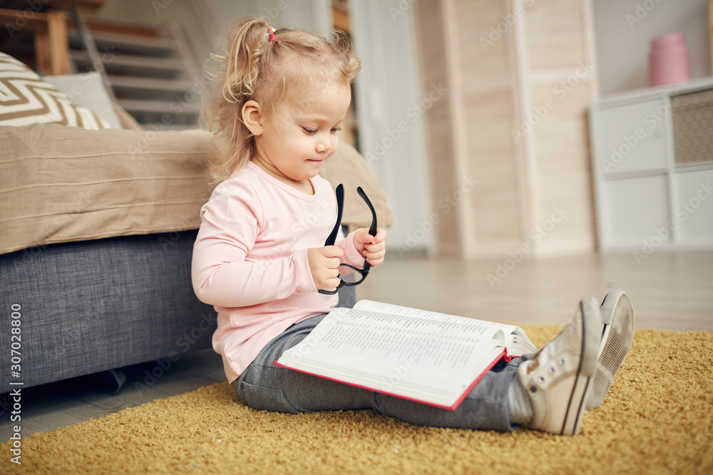 Full length portrait of cute little girl reading big book and plying with glasses while sitting on floor in cozy home interior, copy space Full length portrait of cute little girl reading big book and plying with glasses while sitting on floor in cozy home interior, copy space