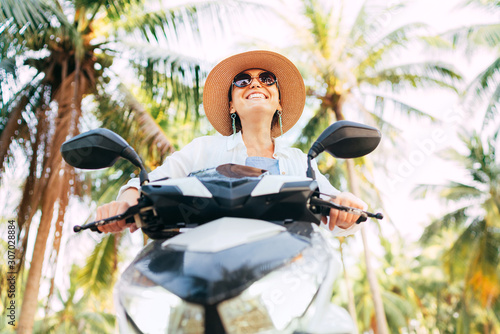 Happy smiling woman in straw hat and sunglasses riding motorbike under palm tree.. Careless vacation time concept image.