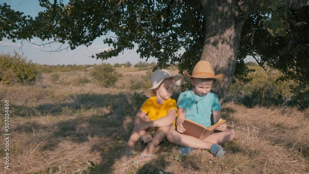 A boy and a girl are reading under a tree. An older brother reads to his sister on a hot sunny day. Summer vacation. Happy childhood. Best childhood friends. A boy and a girl are reading under a tree. An older brother reads to his sister on a hot sunny day. Summer vacation. Happy childhood. Best childhood friends.