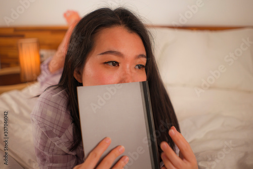 young beautiful and sweet Asian Chinese woman in bed reading book novel feeling happy and playful or studying at home wearing cute pajamas looking cozy and homey