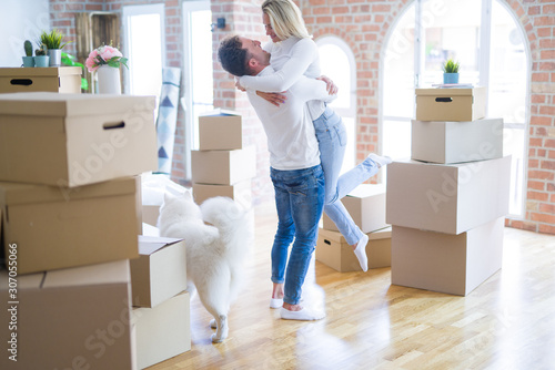 Young beautiful couple with dog hugging at new home around cardboard boxes