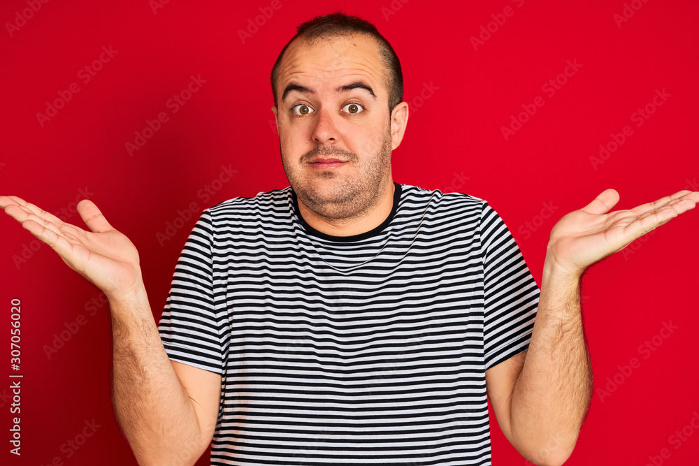 Young man wearing striped navy t-shirt standing over isolated red background clueless and confused expression with arms and hands raised. Doubt concept. Young man wearing striped navy t-shirt standing over isolated red background clueless and confused expression with arms and hands raised. Doubt concept.