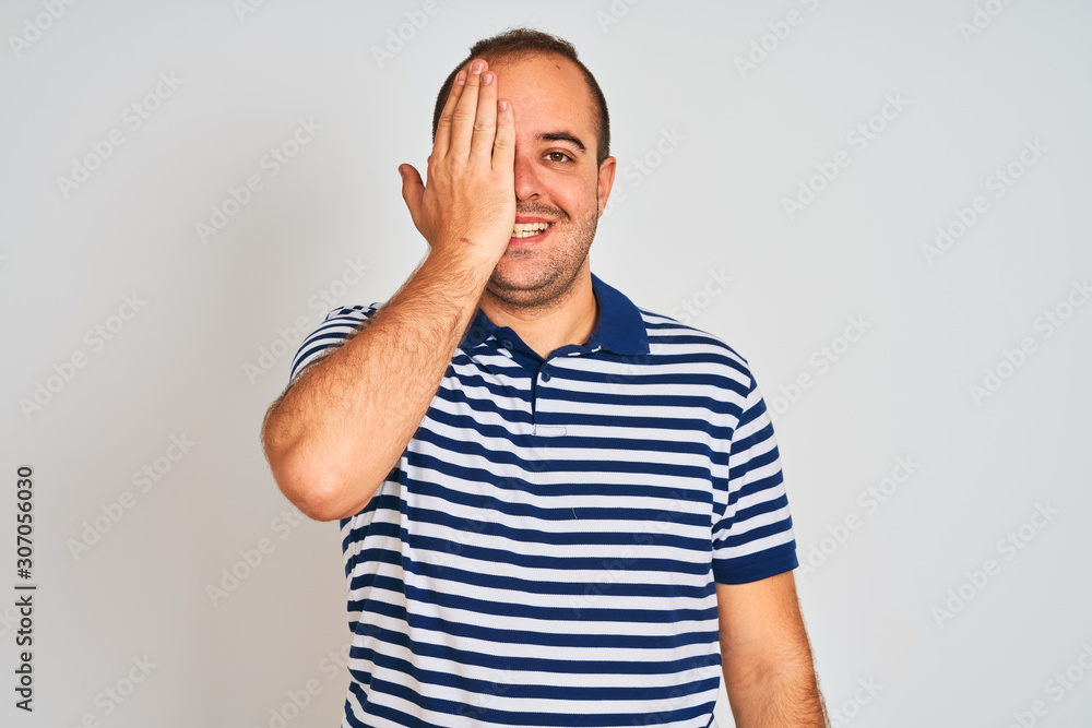 Young man wearing casual striped polo standing over isolated white background covering one eye with hand, confident smile on face and surprise emotion. Young man wearing casual striped polo standing over isolated white background covering one eye with hand, confident smile on face and surprise emotion.