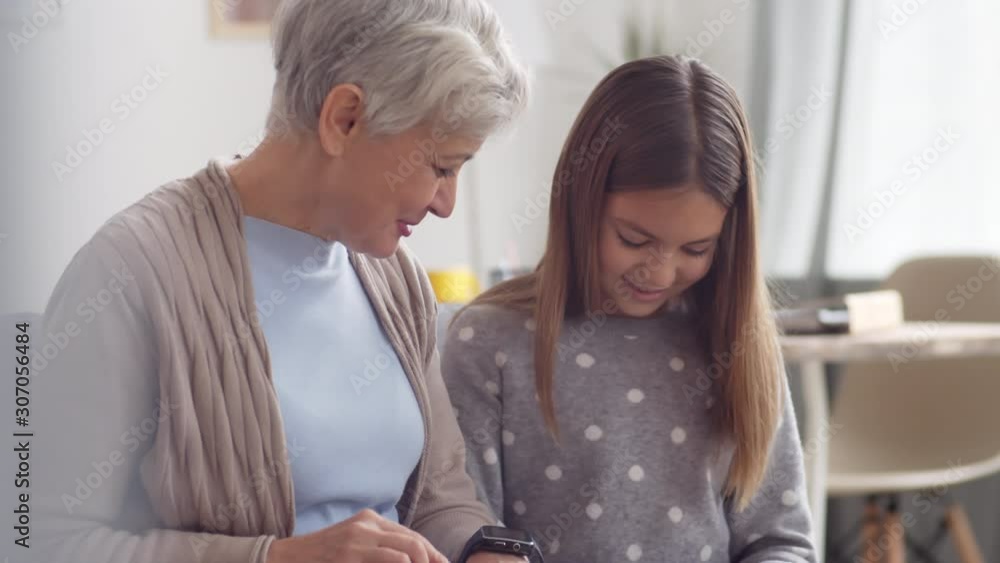 Medium shot of senior active Caucasian woman with grey hair and smart watch on her wrist discussing modern gadgets usage with her lovely granddaughter sitting nearby on sofa Medium shot of senior active Caucasian woman with grey hair and smart watch on her wrist discussing modern gadgets usage with her lovely granddaughter sitting nearby on sofa