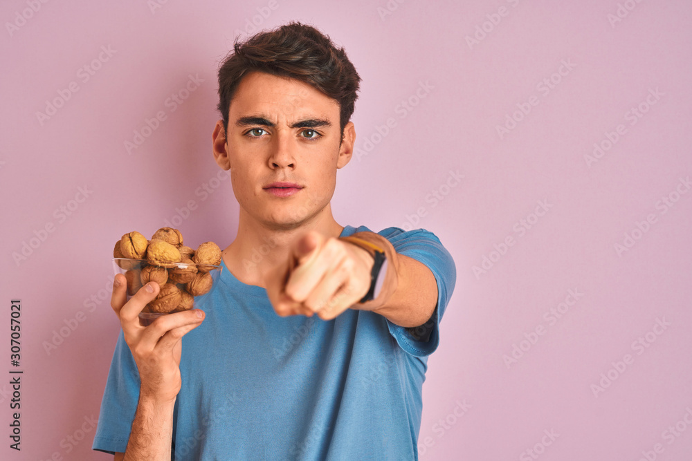 Teenager boy holding bunch of natural walnuts over pink isolated background pointing with finger to the camera and to you, hand sign, positive and confident gesture from the front