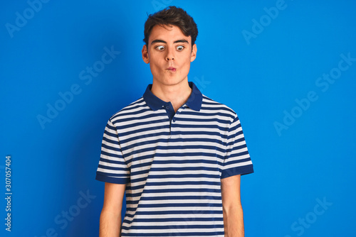 Teenager boy wearing casual t-shirt standing over blue isolated background making fish face with lips, crazy and comical gesture. Funny expression.
