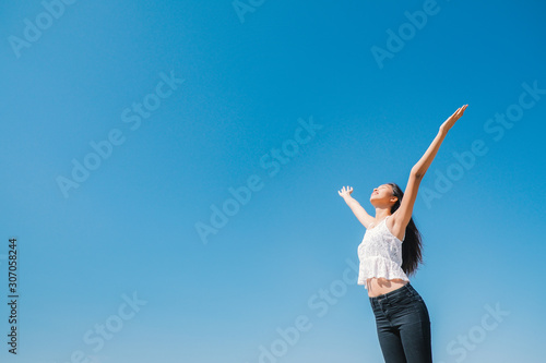 Happy woman with tanned slim body breathing fresh air raising her arms up, enjoying a sunny summer holiday on beach destination against blue sky, outdoors. Travel and well being lifestyle.