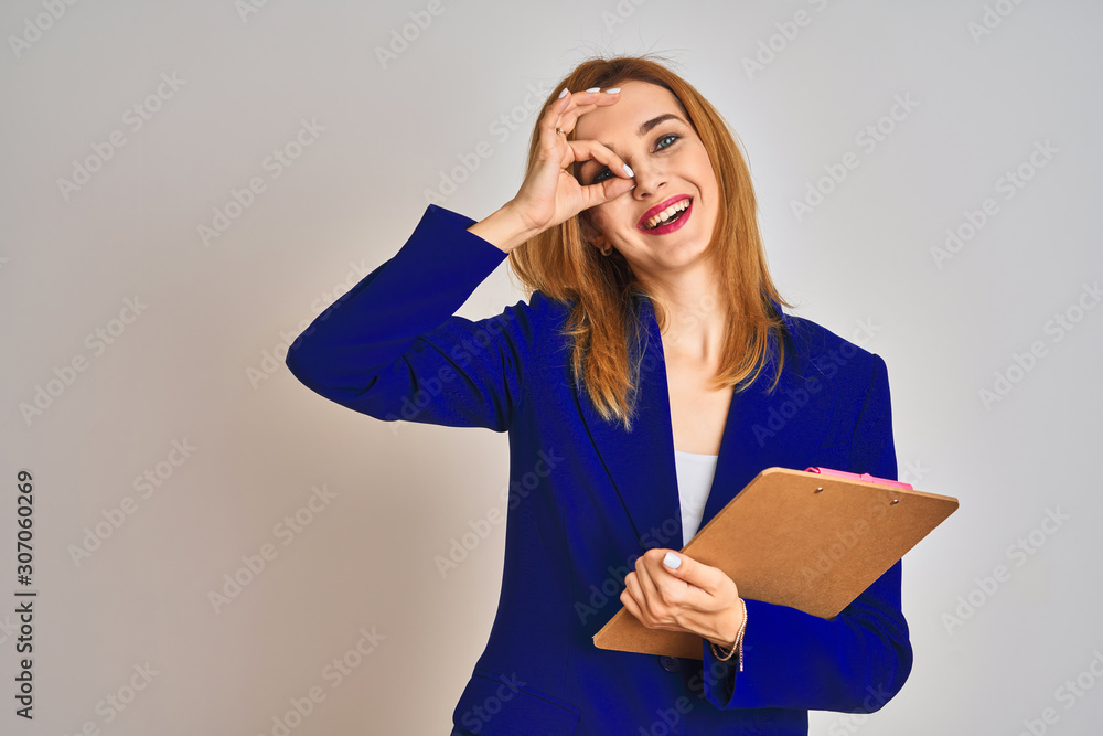 Redhead business caucasian woman holding clipboard over isolated background with happy face smiling doing ok sign with hand on eye looking through fingers Redhead business caucasian woman holding clipboard over isolated background with happy face smiling doing ok sign with hand on eye looking through fingers