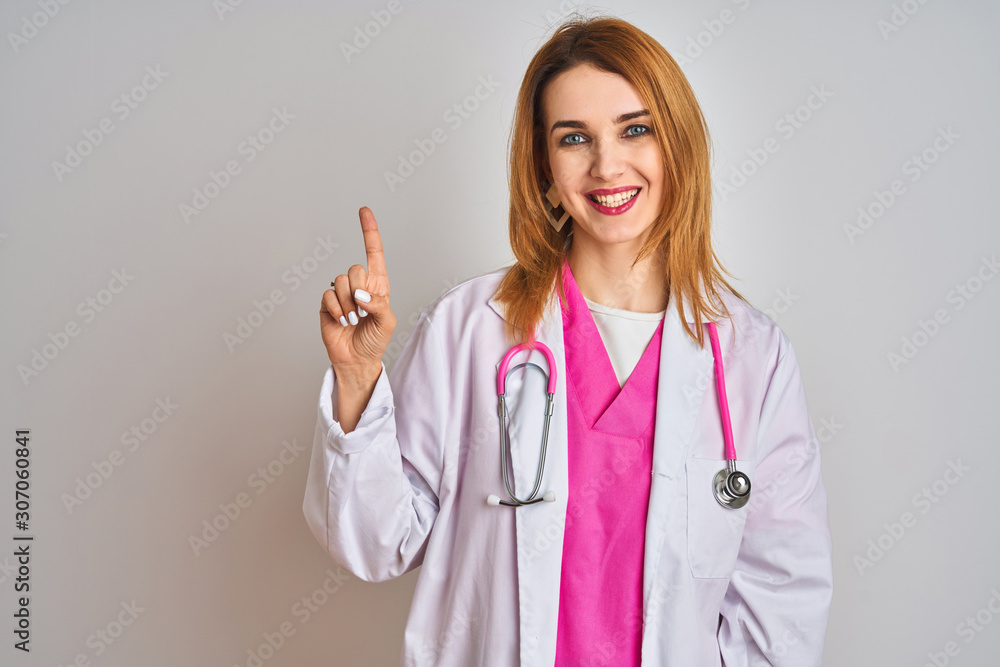 Redhead caucasian doctor woman wearing pink stethoscope over isolated background showing and pointing up with finger number one while smiling confident and happy. Redhead caucasian doctor woman wearing pink stethoscope over isolated background showing and pointing up with finger number one while smiling confident and happy.