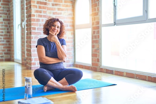 Middle age beautiful sportswoman wearing sportswear sitting on mat practicing yoga at home looking confident at the camera with smile with crossed arms and hand raised on chin. Thinking positive.