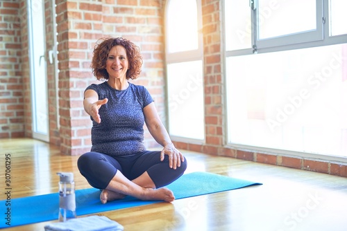 Middle age beautiful sportswoman wearing sportswear sitting on mat practicing yoga at home smiling friendly offering handshake as greeting and welcoming. Successful business.