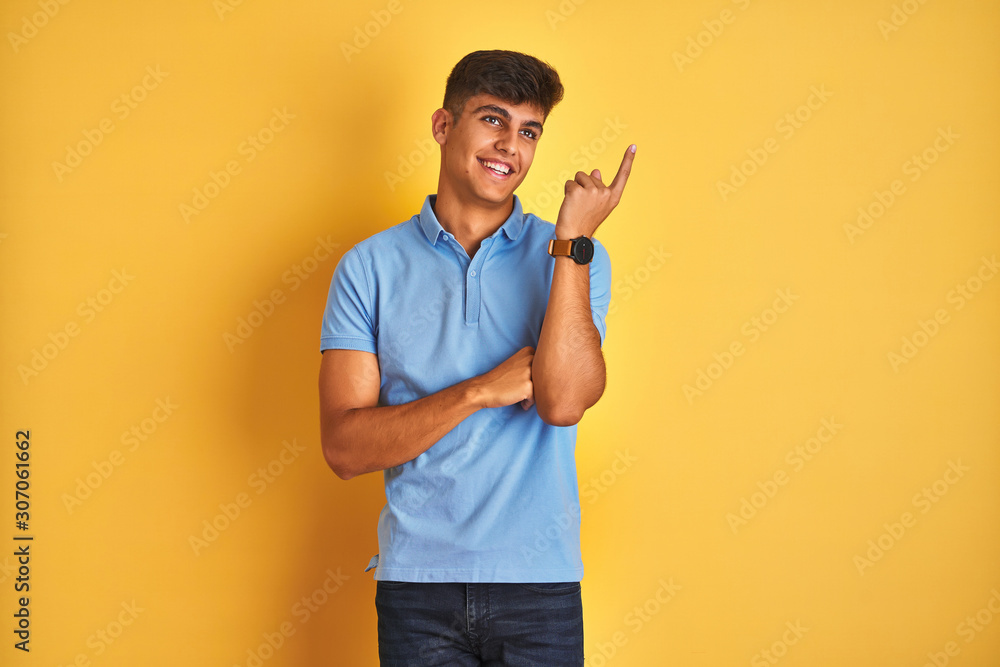 Young indian man wearing blue polo standing over isolated yellow background with a big smile on face, pointing with hand and finger to the side looking at the camera. Young indian man wearing blue polo standing over isolated yellow background with a big smile on face, pointing with hand and finger to the side looking at the camera.
