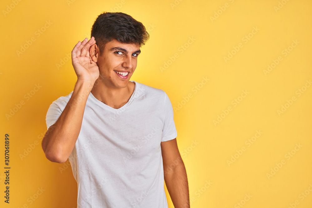 Young indian man wearing white t-shirt standing over isolated yellow background smiling with hand over ear listening an hearing to rumor or gossip. Deafness concept. Young indian man wearing white t-shirt standing over isolated yellow background smiling with hand over ear listening an hearing to rumor or gossip. Deafness concept.
