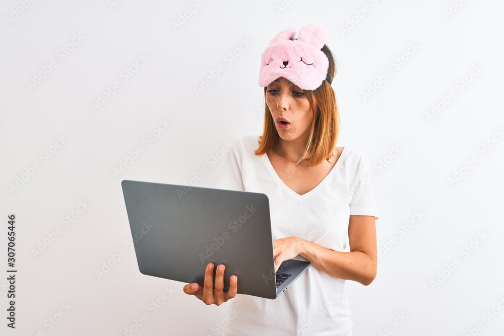 Beautiful redhead woman wearing sleeping mask using laptop over isolated background scared in shock with a surprise face, afraid and excited with fear expression Beautiful redhead woman wearing sleeping mask using laptop over isolated background scared in shock with a surprise face, afraid and excited with fear expression