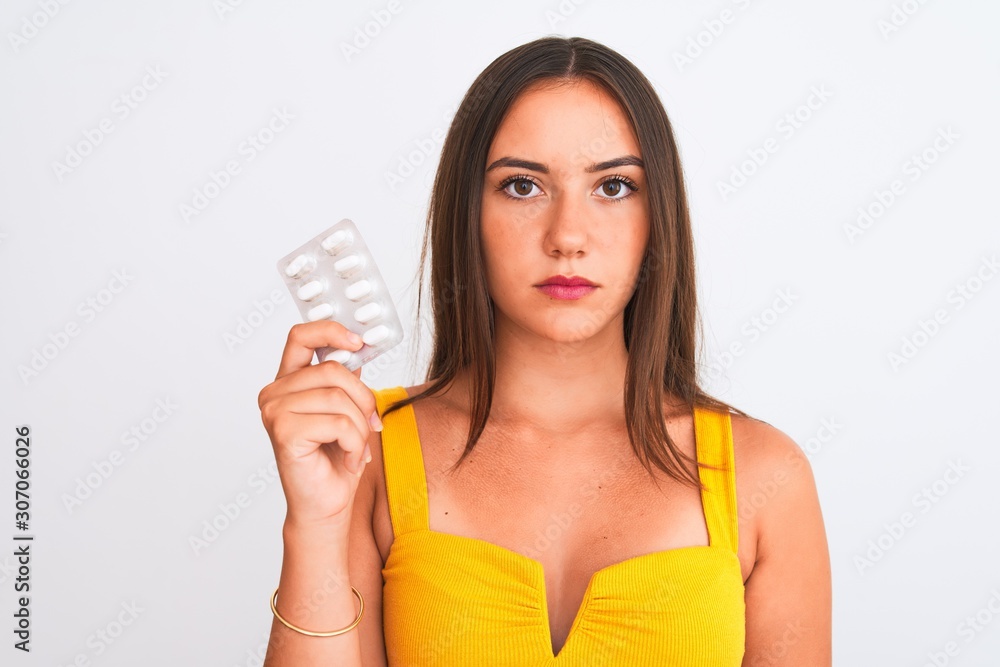 Young beautiful girl holding medicine pills standing over isolated white background with a confident expression on smart face thinking serious Young beautiful girl holding medicine pills standing over isolated white background with a confident expression on smart face thinking serious