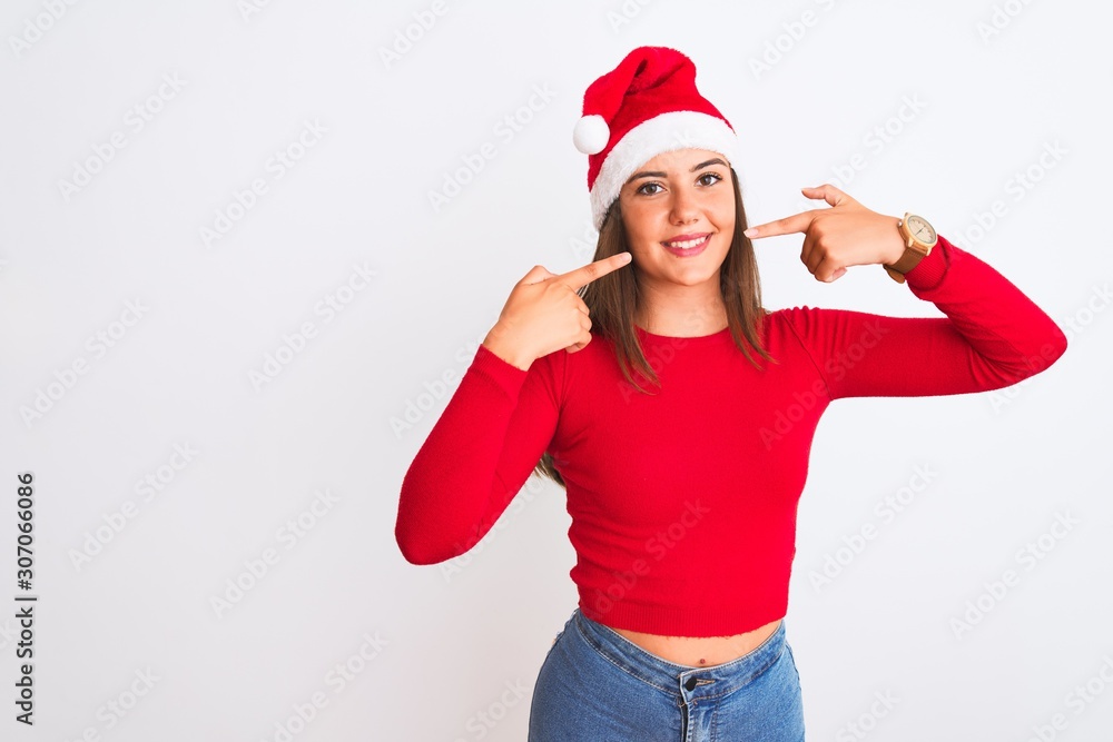 Young beautiful girl wearing Christmas Santa hat standing over isolated white background smiling cheerful showing and pointing with fingers teeth and mouth. Dental health concept. Young beautiful girl wearing Christmas Santa hat standing over isolated white background smiling cheerful showing and pointing with fingers teeth and mouth. Dental health concept.