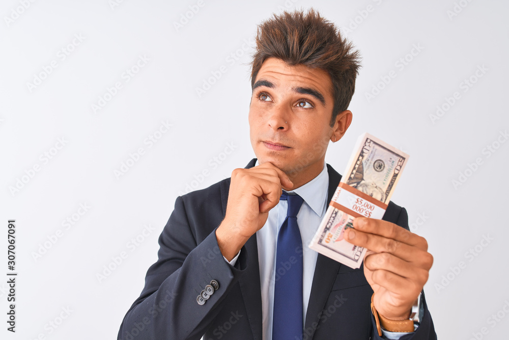 Young handsome businessman wearing suit holding dollars over isolated white background serious face thinking about question, very confused idea