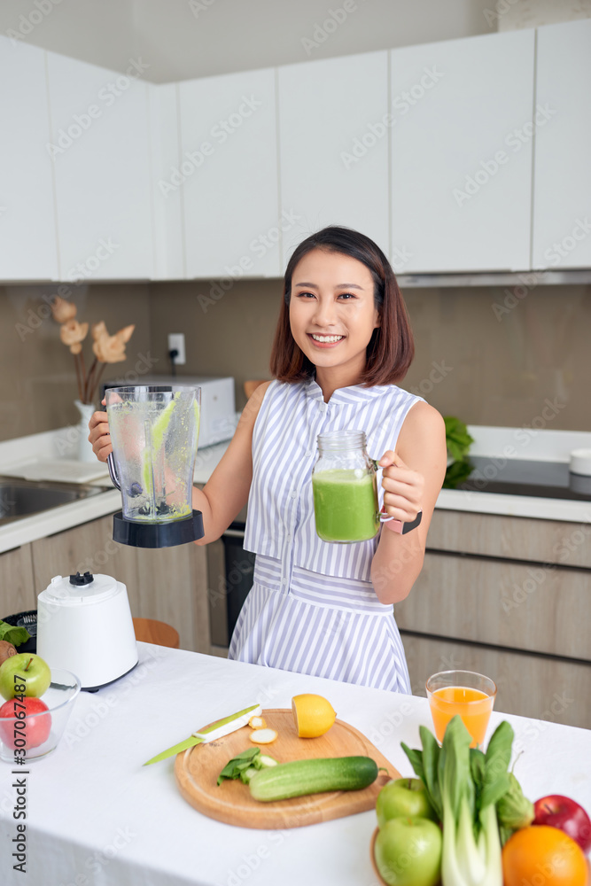 Smiling young Asian woman holding green smoothie in the kitchen at home.