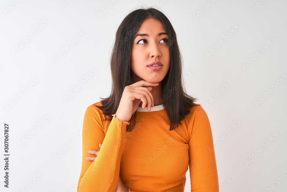 Young beautiful chinese woman wearing orange t-shirt standing over isolated white background with hand on chin thinking about question, pensive expression. Smiling with thoughtful face. Doubt concept. Young beautiful chinese woman wearing orange t-shirt standing over isolated white background with hand on chin thinking about question, pensive expression. Smiling with thoughtful face. Doubt concept.