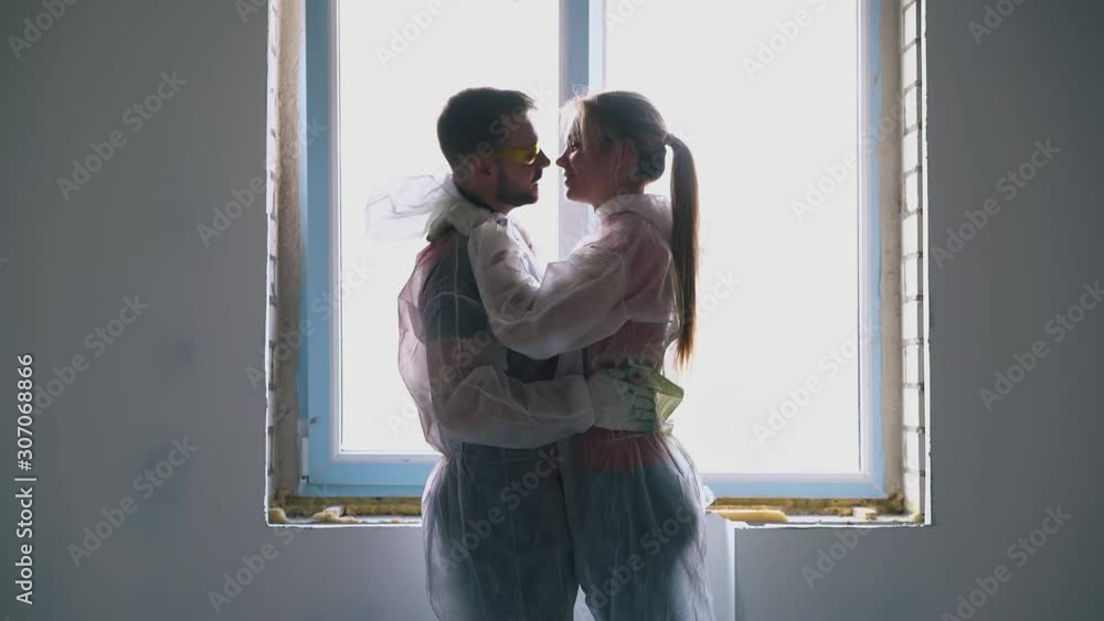 positive young family of happy girl and bearded guy in transparent coveralls dances hugging near window in spacious room positive young family of happy girl and bearded guy in transparent coveralls dances hugging near window in spacious room
