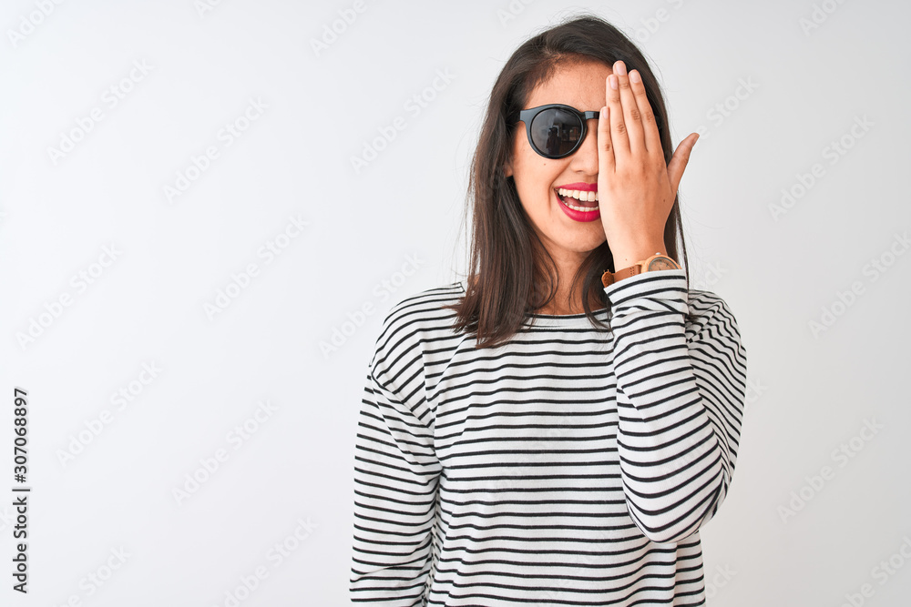 Chinese woman wearing striped t-shirt and sunglasses standing over isolated white background covering one eye with hand, confident smile on face and surprise emotion. Chinese woman wearing striped t-shirt and sunglasses standing over isolated white background covering one eye with hand, confident smile on face and surprise emotion.