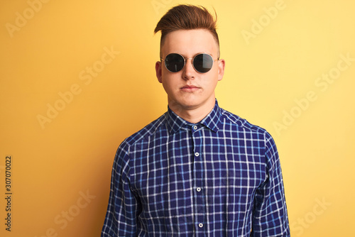 Young handsome man wearing casual shirt and sunglasses over isolated yellow background with serious expression on face. Simple and natural looking at the camera. © Krakenimages.com