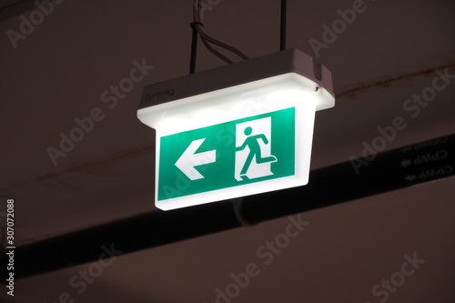 Glowing Emergency exit sign with left arrow at a building. Safety first concept. Copy space wallpaper. Exit signs with light in a parking of building.