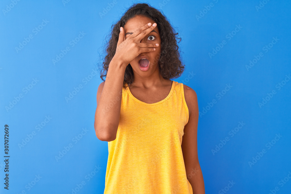 Young brazilian woman wearing yellow t-shirt standing over isolated blue background peeking in shock covering face and eyes with hand, looking through fingers with embarrassed expression. Young brazilian woman wearing yellow t-shirt standing over isolated blue background peeking in shock covering face and eyes with hand, looking through fingers with embarrassed expression.