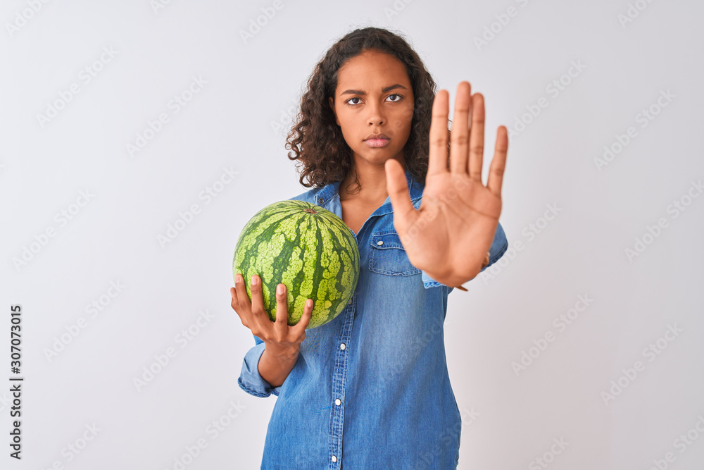 Young brazilian woman holding watermelon standing over isolated white background with open hand doing stop sign with serious and confident expression, defense gesture Young brazilian woman holding watermelon standing over isolated white background with open hand doing stop sign with serious and confident expression, defense gesture
