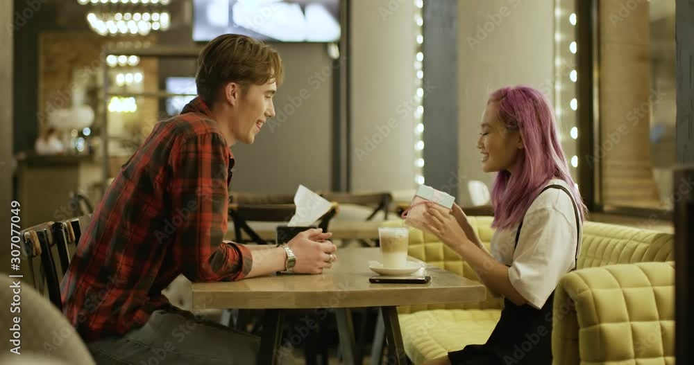 Young couple in cafe, evening in the city, guy gives a gift to the asian girl, positive emotions, blurry lights on background, 4k slow motion. Young couple in cafe, evening in the city, guy gives a gift to the asian girl, positive emotions, blurry lights on background, 4k slow motion.