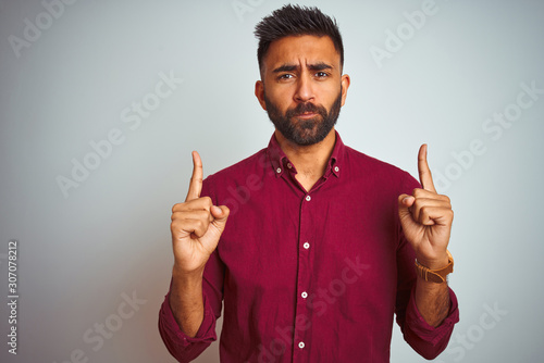 Young indian man wearing red elegant shirt standing over isolated grey background Pointing up looking sad and upset, indicating direction with fingers, unhappy and depressed.