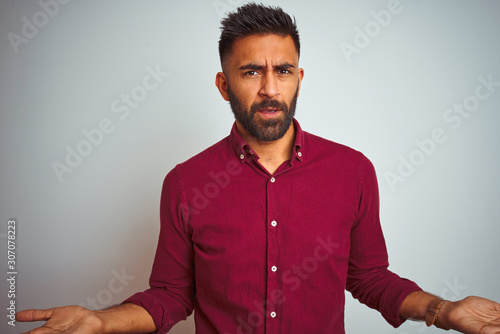 Young indian man wearing red elegant shirt standing over isolated grey background clueless and confused with open arms, no idea concept.
